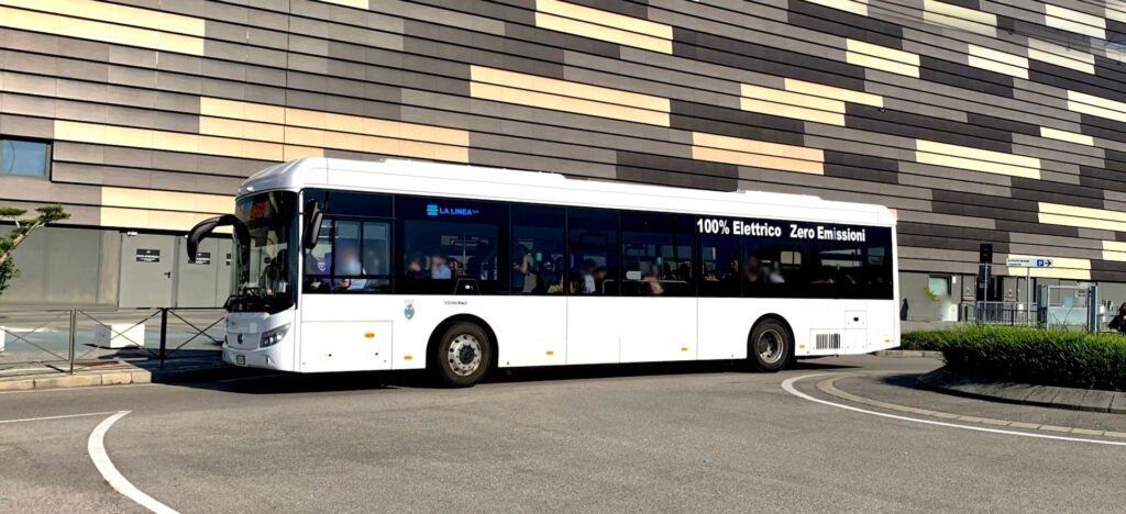 Modern white electric bus displaying "100% Elettrico Zero Emissioni" on its side, parked near a contemporary building with geometric tile facade in an urban setting.