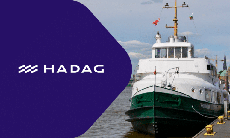 HADAG ferry boat docked in Hamburg with white superstructure and green hull, featuring company branding and maritime flags against cloudy sky.