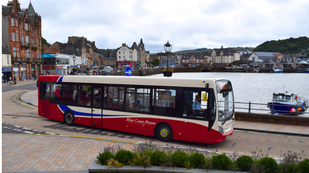 Red and cream tour bus parked along a Scottish waterfront promenade with historic stone buildings and boats visible across the harbor.