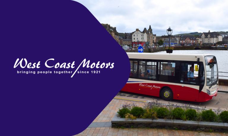 West Coast Motors red and white bus parked by a scenic Scottish waterfront with historic buildings and a blue directional sign visible in the background.