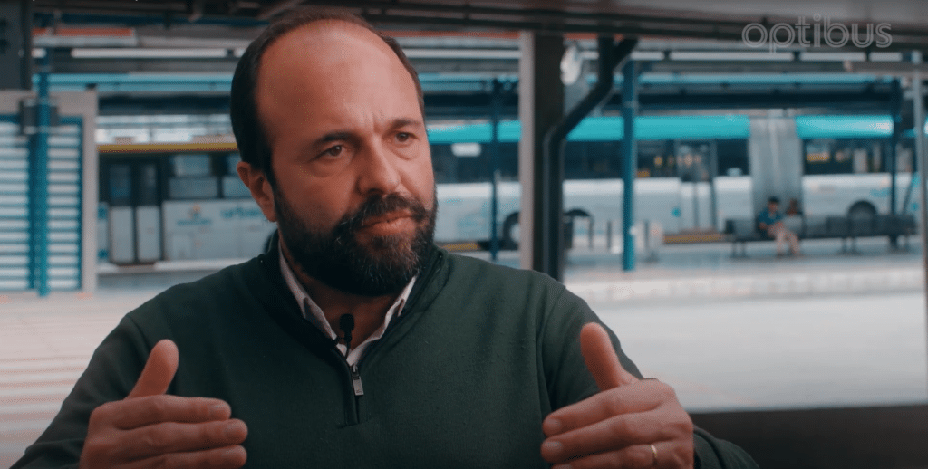 A bearded man in a dark green zip-up jacket speaks expressively with open hands at what appears to be a modern transit station or airport terminal with turquoise architectural elements.