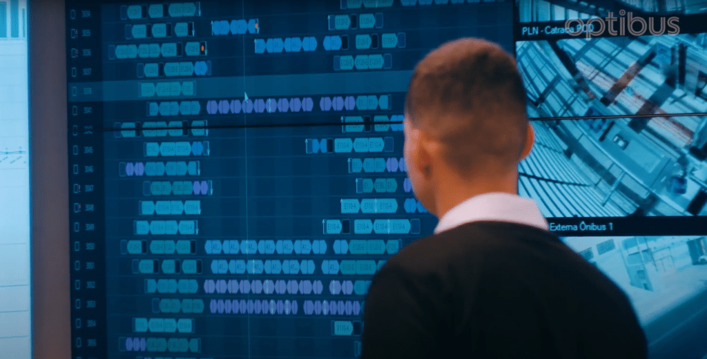 Professional observes digital monitoring system displaying complex data visualizations and network infrastructure on large blue-tinted display screens in control room environment.