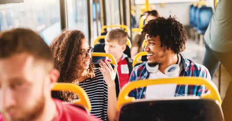Diverse passengers enjoy casual conversation on a public bus, with a woman and man chatting happily while other riders sit nearby in the modern transit vehicle.