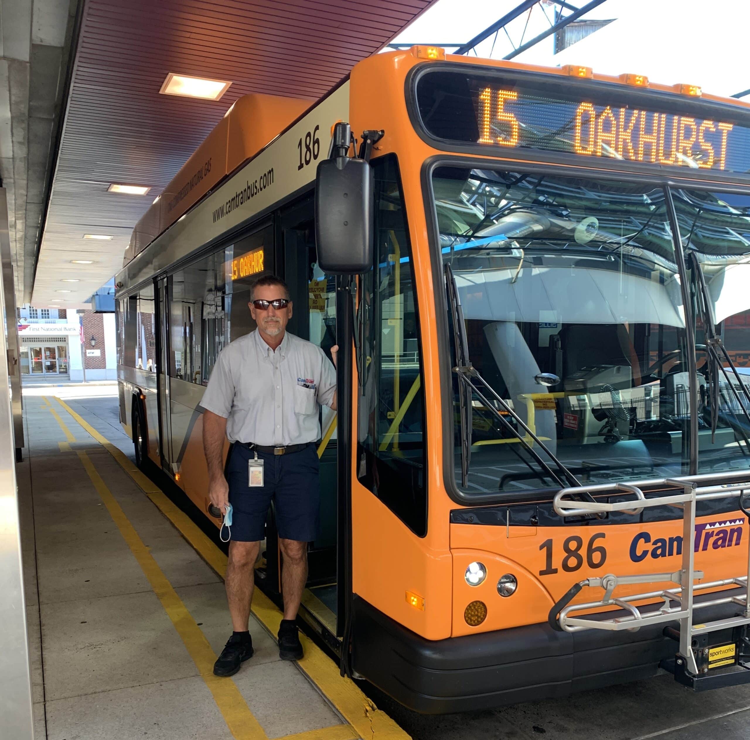 Bus driver stands in front of orange Route 15 Orkhurst transit bus at station platform during daytime service.