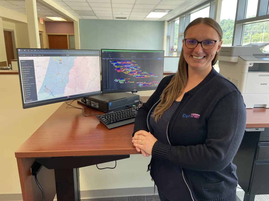 Professional woman at CamTran workstation displays dual monitors showing geographical mapping and data analytics software in modern office setting.