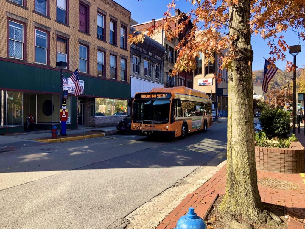 Orange downtown shuttle bus travels through a charming autumn street lined with colorful fall foliage, brick buildings, and American flags on a clear day.