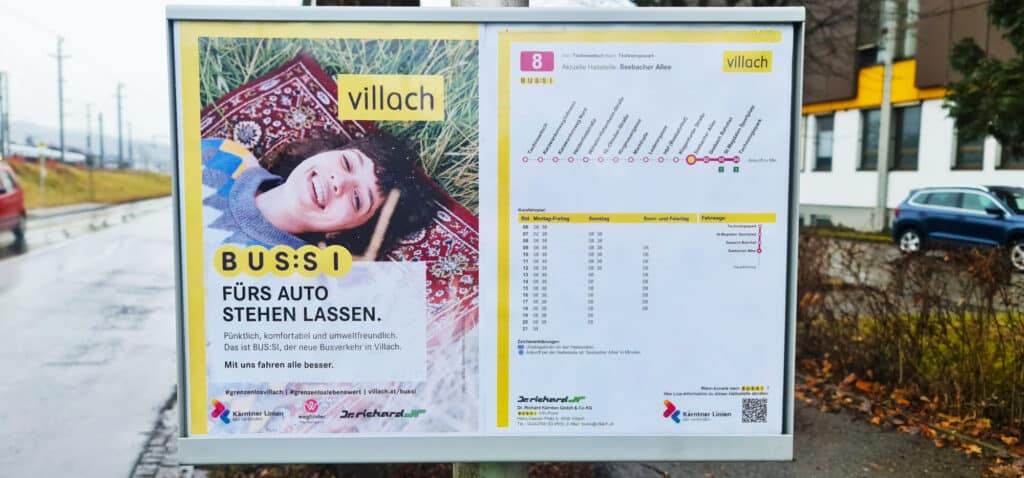 Bus shelter advertisement for Villach's BUS:SI transit system featuring a smiling child and route schedule information for the Seebacher Allee bus line.