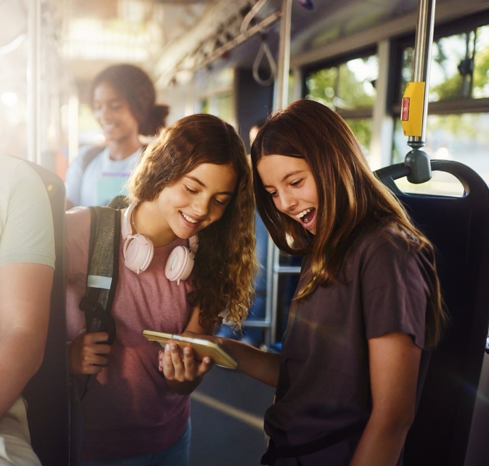 Two teenage girls on a bus smile while looking at a tablet together, wearing headphones around their necks. Sunlight streams through the windows as other passengers sit in the background.