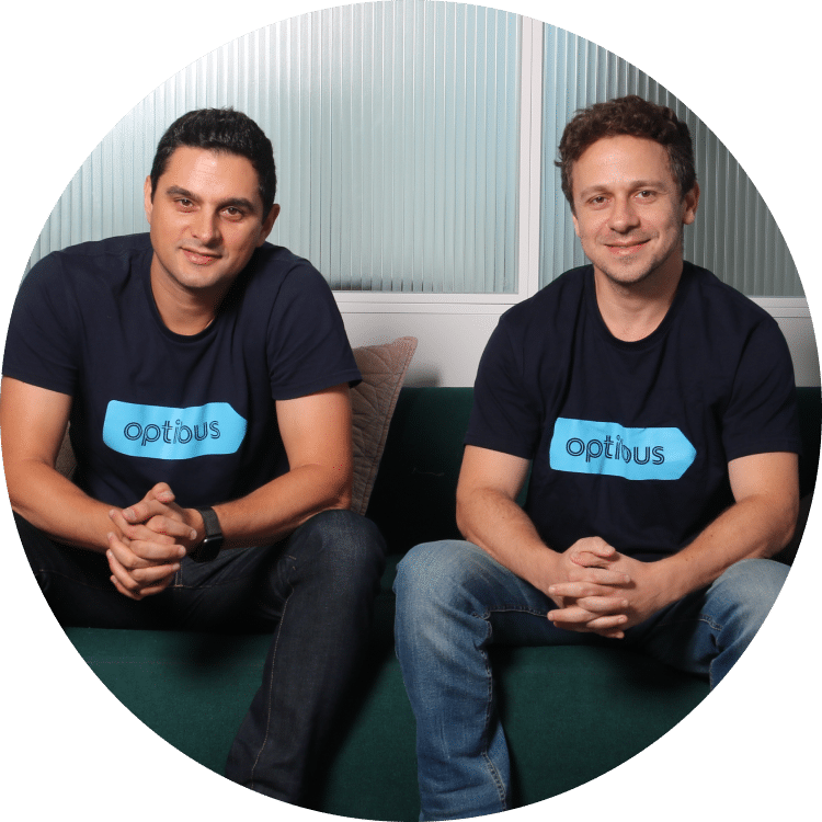 Two men wearing black Optibus branded t-shirts sit together on a green bench against a modern corrugated metal wall background in a professional team photo.