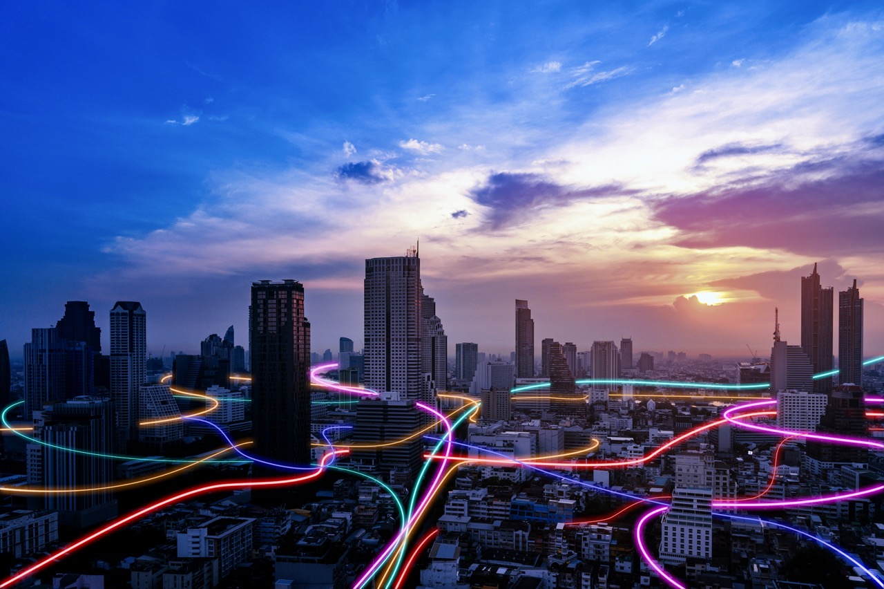 Neon light trails weave through a futuristic cityscape at sunset, with illuminated buildings silhouetted against a vibrant pink and purple sky. The glowing paths of magenta, cyan, and gold