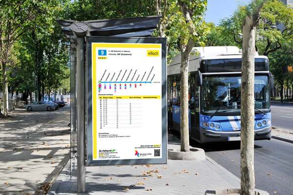 Public bus stop shelter with yellow transit information display and schedule chart, featuring blue city buses stopped on a tree-lined urban street.