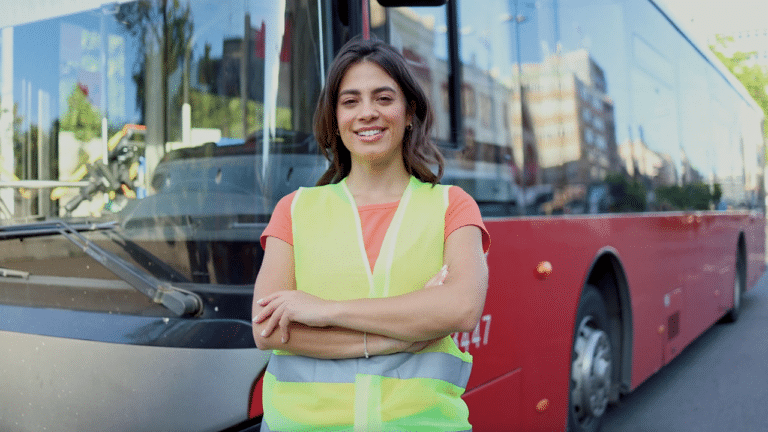 Professional transit worker in safety vest stands confidently in front of buses with city skyline reflected behind her.