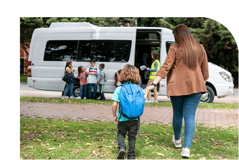 Teacher guides children with backpacks toward a white van during a school field trip outdoors. Multiple kids in casual clothing gather near the vehicle on a grassy area.