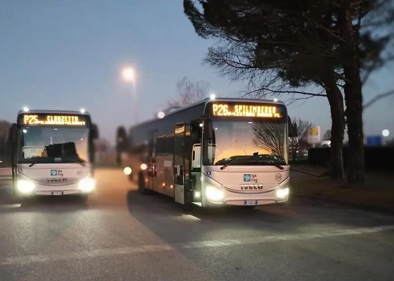 Public transit buses parked at dusk with illuminated route displays, featuring Iveco vehicles with headlights on during evening hours in an urban setting.