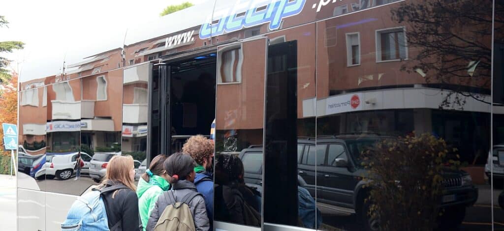 Passengers board a long-distance coach bus at a street stop, with the vehicle's open door and several people waiting to enter the transport service.