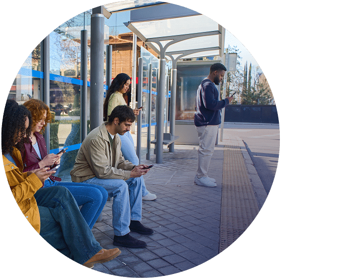 People wait at a modern bus shelter on a sunny day. Several passengers sit on benches while others stand, checking phones or looking down the street.