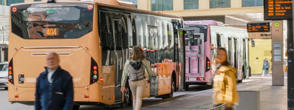 Busy urban bus station with multiple orange and pink buses lined up, passengers boarding and walking between vehicles on a sunny day.