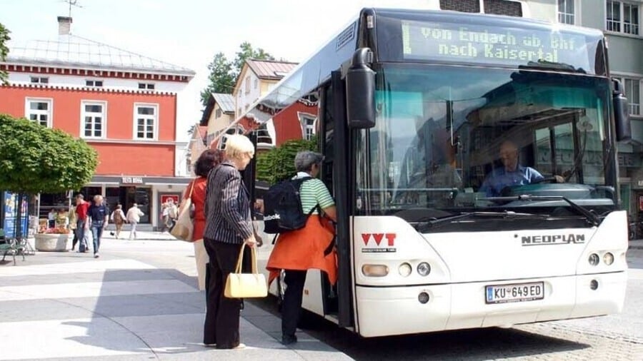 Public bus stops in a European town square where elderly passengers board near a red historic building and traditional architecture.