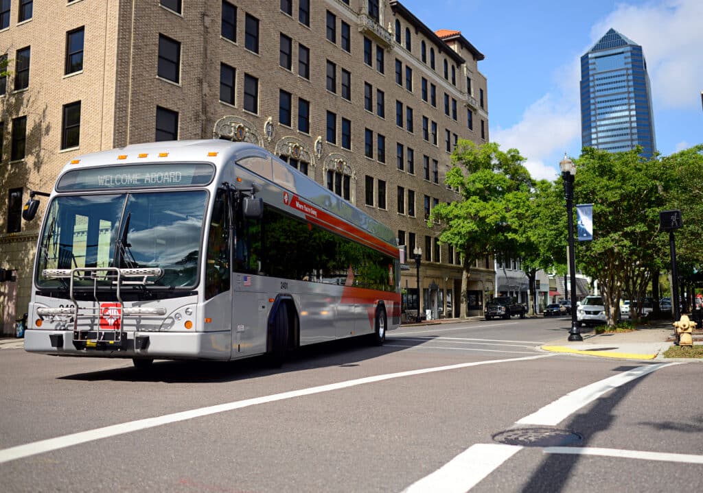 Public transit bus displaying "Welcome Aboard" on an urban street lined with historic brick buildings and modern skyscrapers under clear blue sky.