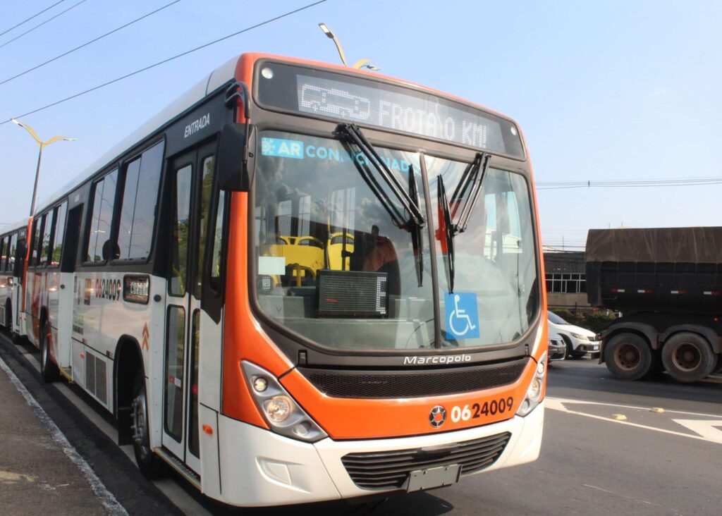 Orange and white Marcopolo bus with accessibility features and electronic destination display parked on a city street next to commercial vehicles and buildings.