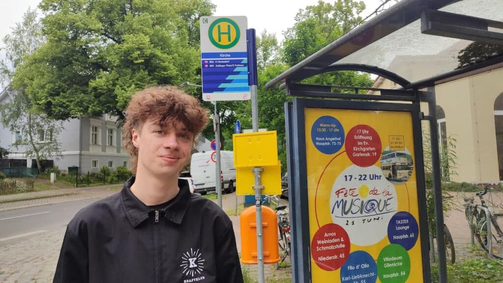 Young man with curly brown hair at a German bus stop, standing beside a yellow advertisement shelter with event listings and transit information visible in the background.