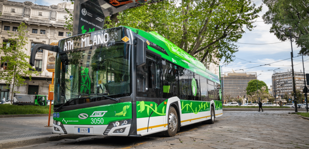 Green and white ATM Milano bus number 3050 travels through a Milan city square, featuring eco-friendly design with distinctive artwork and yellow accents on its exterior.