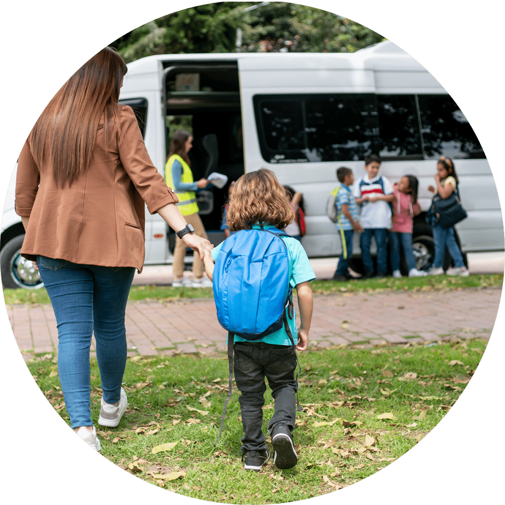 A woman guides a young child wearing a blue backpack toward a school bus with other children nearby. The scene captures a typical school drop-off moment on a grassy outdoor area.