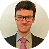 Professional headshot of a young man wearing glasses and a burgundy tie against a neutral beige background. He has a friendly expression suitable for business or corporate use.