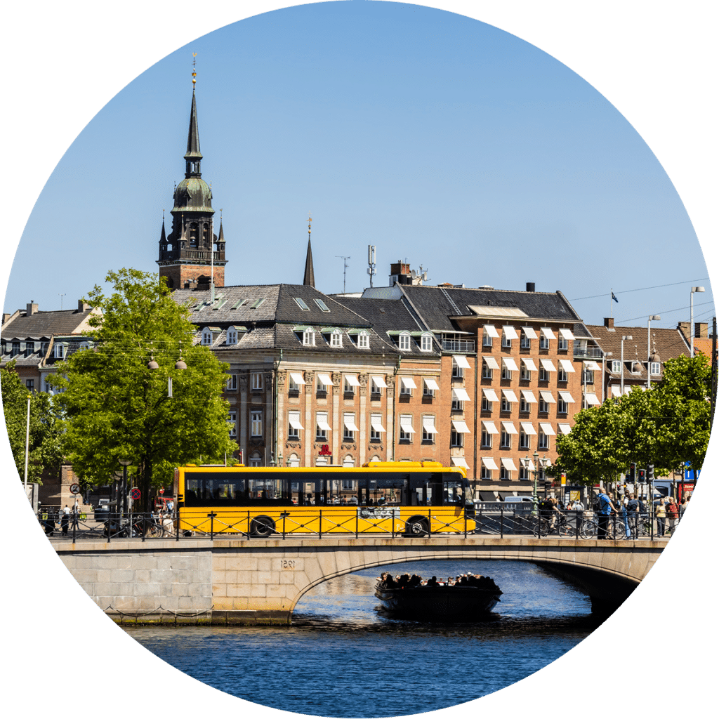 Yellow tram crosses a stone bridge over Copenhagen's canal, with historic brick buildings and a distinctive church tower rising behind it under clear blue skies.