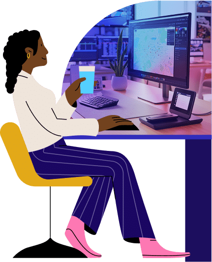 A woman sits at a desk working on a computer in a modern office setup with dual monitors and tech equipment. She wears professional attire and appears focused on her digital design work.