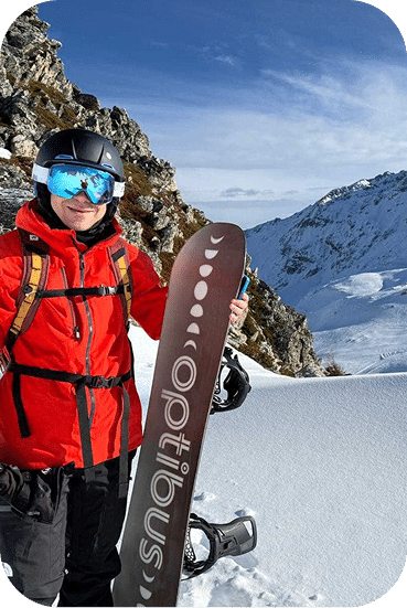 Snowboarder in red jacket and blue goggles holds a brown snowboard on a snowy mountain slope with rocky peaks and valley views in the background.