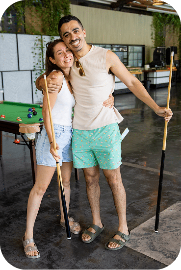 A young couple poses together holding pool cues in a recreational game room, smiling warmly while standing near a billiards table.