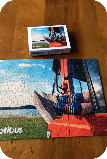 Optibus puzzle box and assembled jigsaw featuring a red agricultural harvester in a field with blue sky and landscape background.