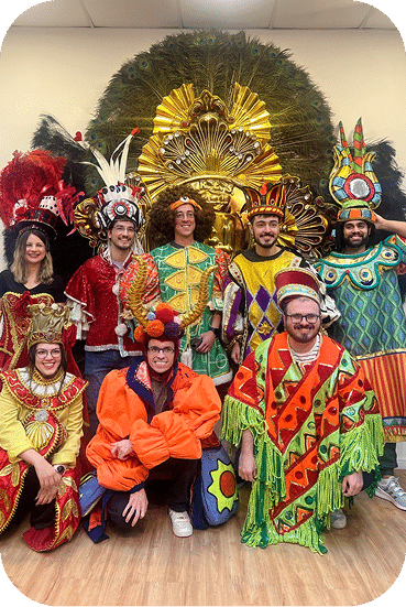 Group of people wearing elaborate colorful carnival costumes and headdresses pose together in front of a large golden decorative backdrop.