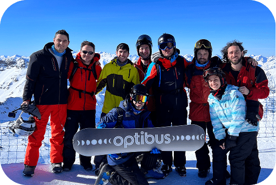 Optibus team celebrates on a snowy mountain slope with skis and snowboards, holding their company banner under clear blue skies with alpine peaks visible.