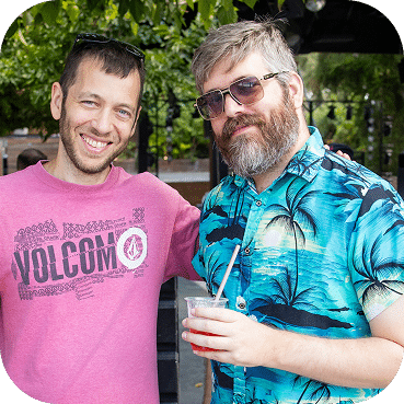 Two men pose outdoors smiling at the camera. The man on the left wears a pink Volcom shirt, while the man on the right sports a blue tropical print shirt and sunglasses.
