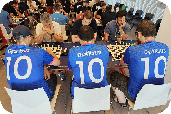 Chess tournament players wearing blue Optibus team jerseys compete at tables during a corporate event with spectators in the background.