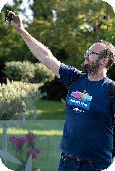 Man in blue shirt takes a selfie outdoors in a garden setting with green foliage and purple flowers visible in the background.