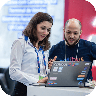 Two professionals wearing conference lanyards collaborate on a laptop at a tech event, with a red branded backdrop visible behind them.