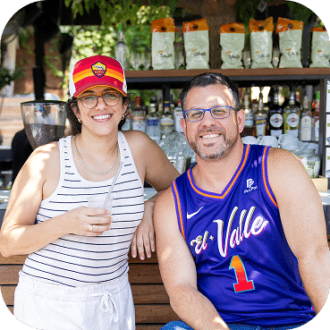 Two people smile at an outdoor bar, one wearing a red cap and striped tank top, the other in a blue basketball jersey. Bottles and plants decorate the wooden counter behind them.