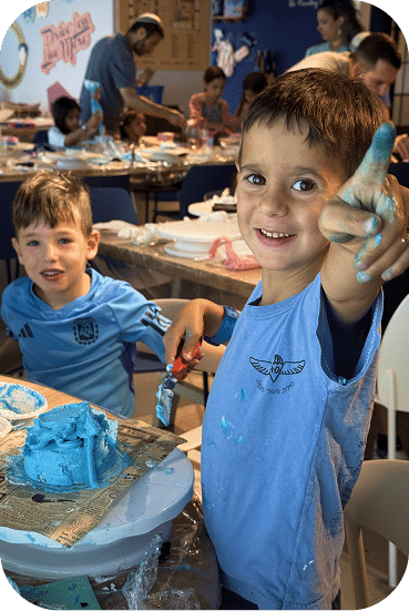 Children in blue shirts enjoy a messy art activity at an indoor event, with one boy proudly displaying his paint-covered hands while smiling at the camera.