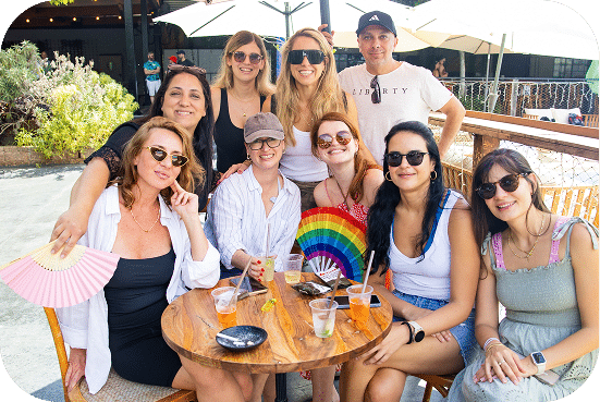 A diverse group of friends poses together outdoors at a sunny venue, smiling while holding a colorful rainbow fan decoration on a wooden table.
