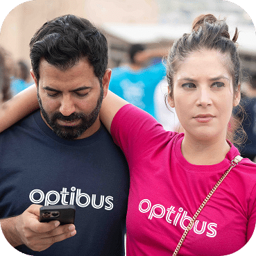 Two people wearing matching Optibus branded shirts stand together at an event, with the man in navy and woman in pink both looking at a mobile device.