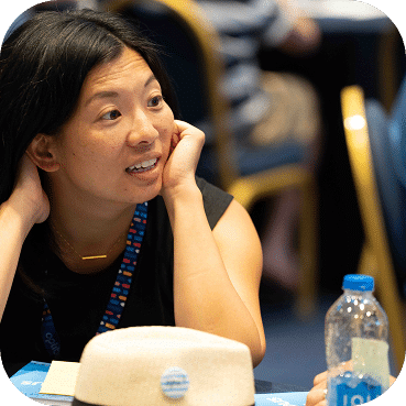 Professional woman with black hair smiles while resting her chin on her hand at a conference table, wearing a blue lanyard beside a water bottle.