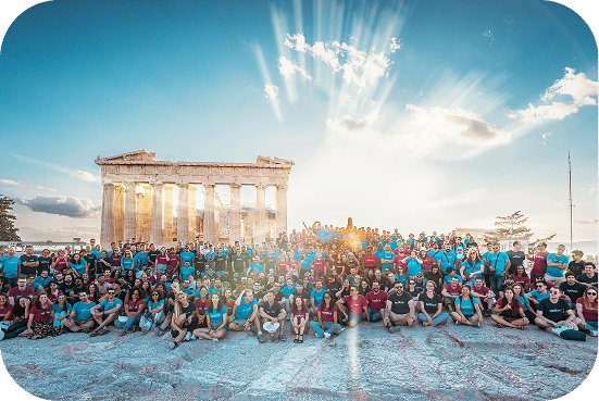 Large group of people wearing blue shirts gathered at the Parthenon in Athens, Greece during golden hour with dramatic sunlight illuminating the ancient temple.