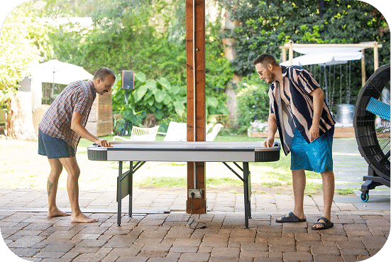 Two men play ping pong at an outdoor table on a brick patio surrounded by lush green trees and garden structures on a sunny day.