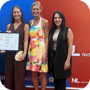 Three women pose together at a professional event against a blue and red backdrop, with the woman on the left holding a certificate award and smiling at the camera.