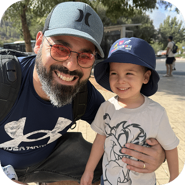 A smiling adult and young child wear baseball caps while posing together outdoors on a sunny day, both displaying genuine joy and warmth in their casual portrait.