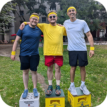 Three men stand on a podium wearing sunglasses and wristbands, celebrating a race or competition victory in an outdoor park setting with historic buildings.