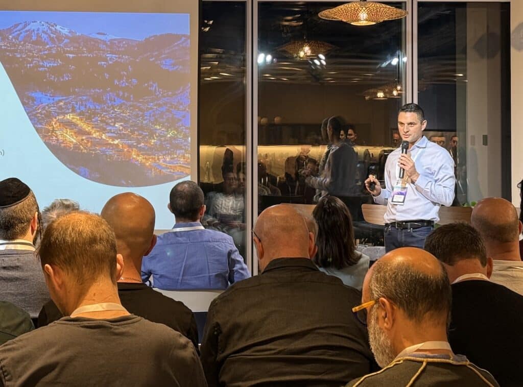A presenter speaks to an audience in a modern venue while displaying a satellite night image of mountainous terrain on a large screen behind him.
